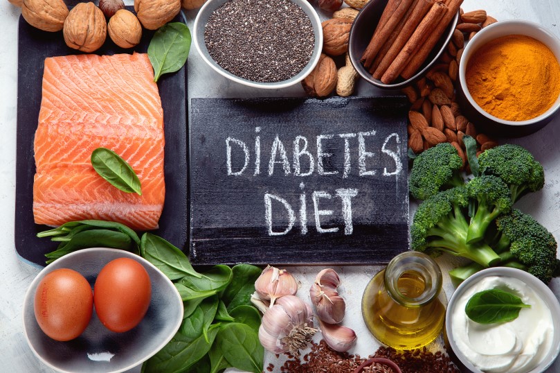 لوحة سوداء مكتوب عليها بالإنكليزية Diabetes diet وحولها خضار متنوعة كالبروكلي والسبانخ بالإضافة للبيض والسمك وزيت الزيتون ومكسرات كاللوز والجوز